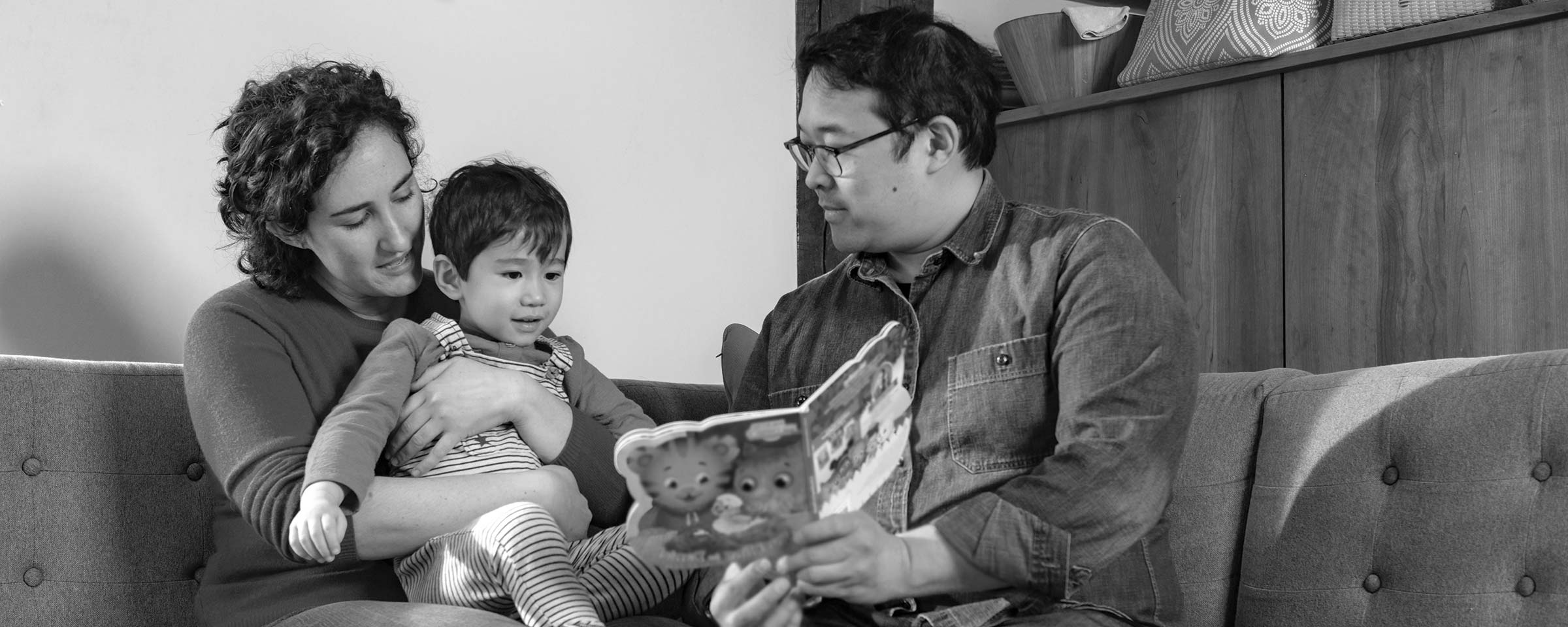 Family of three sitting on their couch in living room with child on lap reading a book together